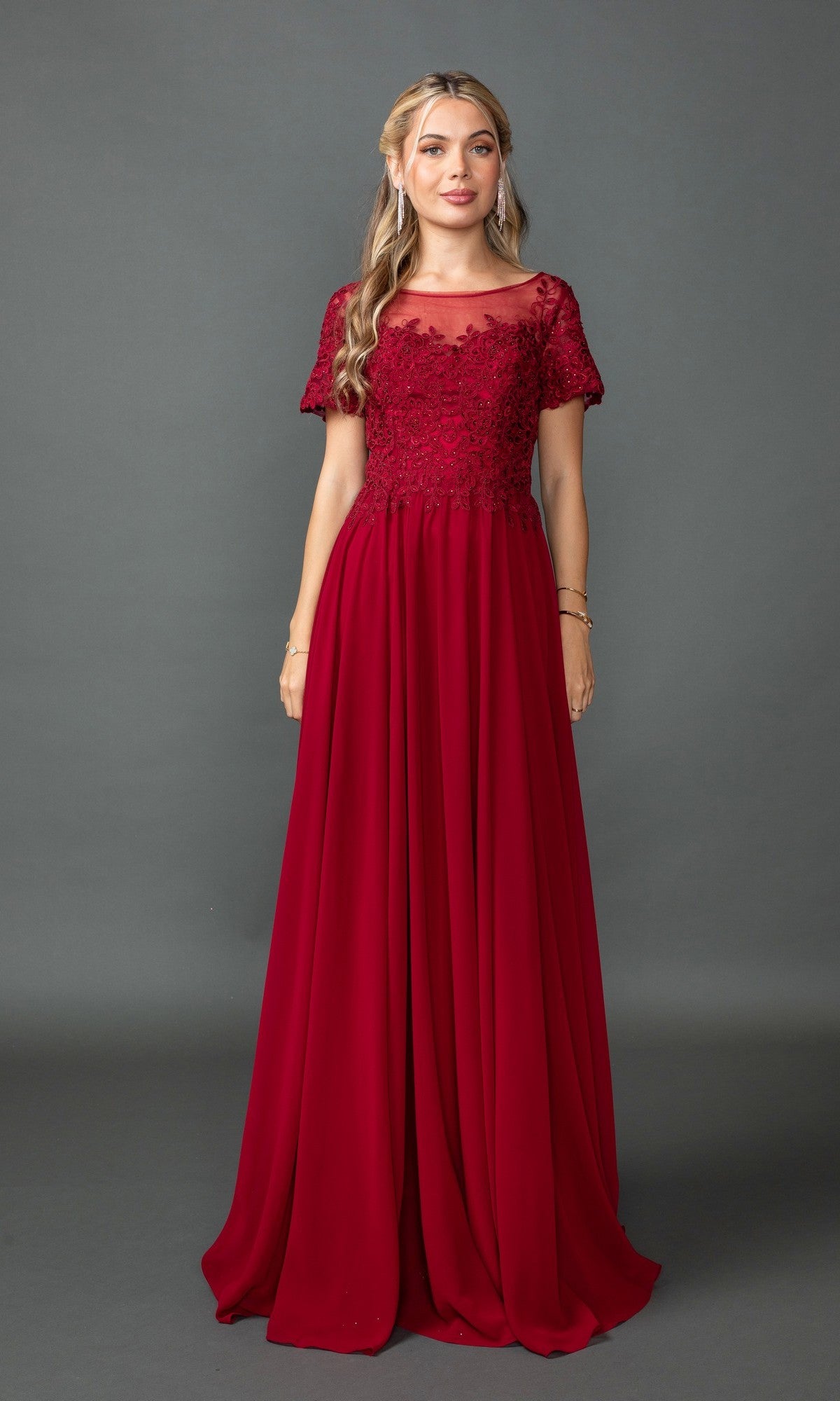 A woman stands against a gray background wearing the Dancing Queen 4433, a short sleeve, floor-length burgundy prom dress with lace and floral details on the bodice.